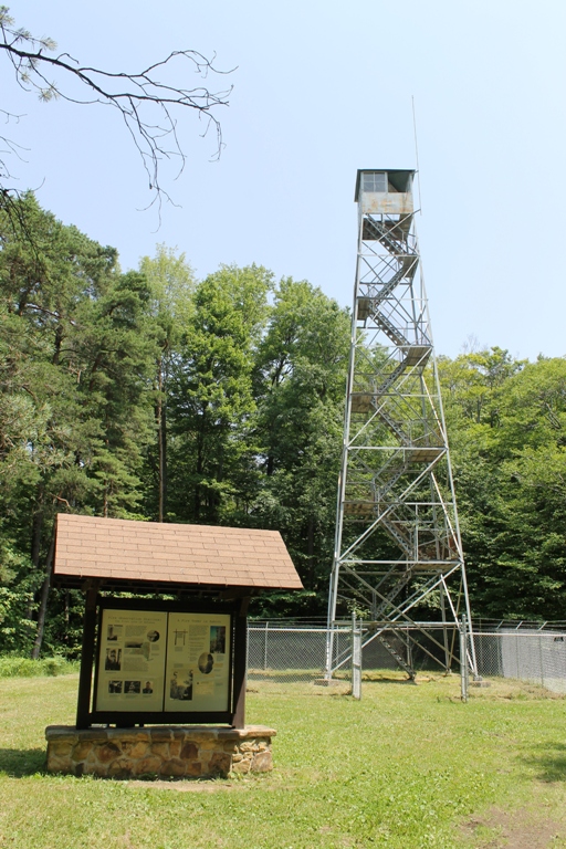 An informational kiosk at the base a fire tower, which is surrounded by a chain link fence.